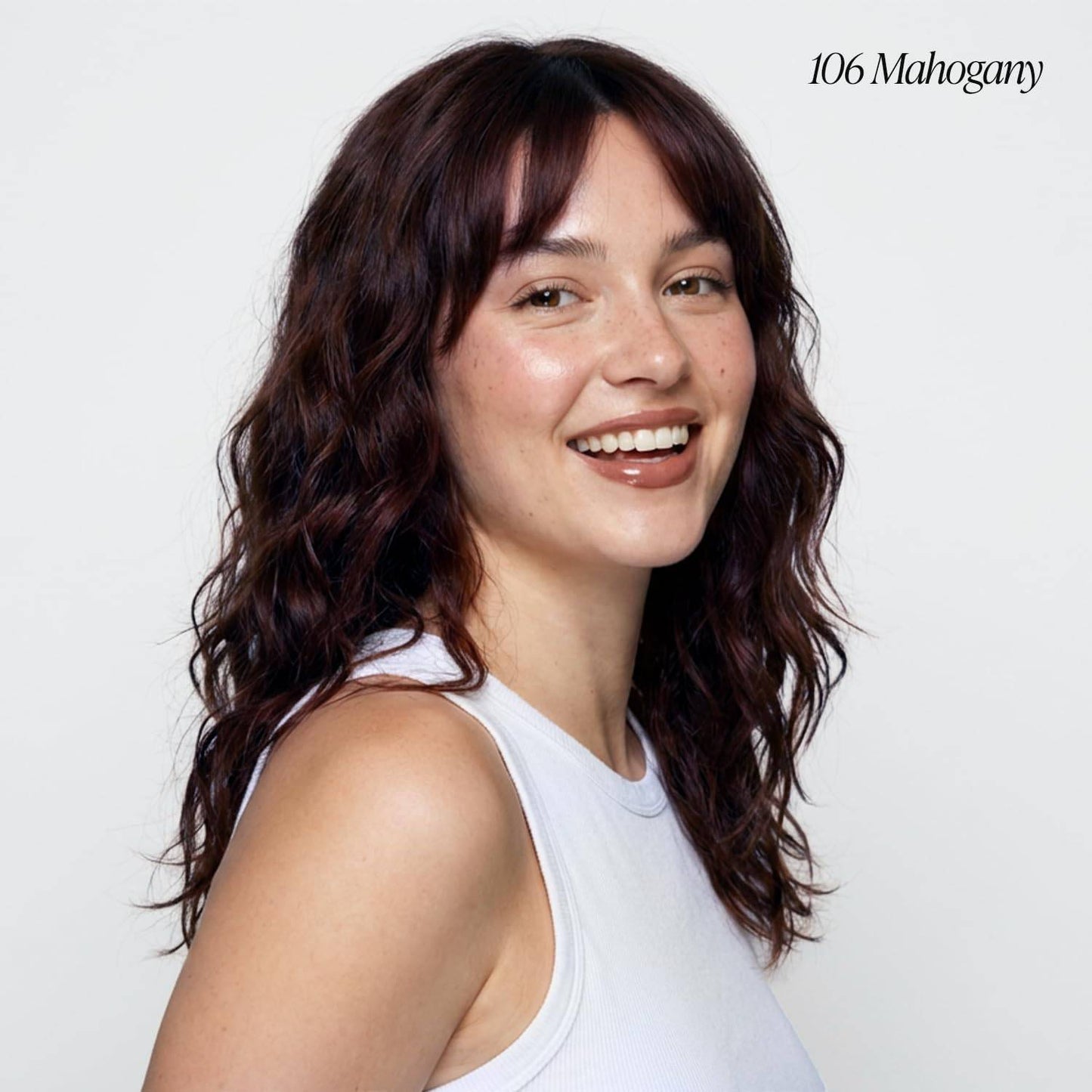 Smiling woman modeling Creative Image Adore #106 Mahogany hair color. She has long, wavy dark reddish-brown hair with bangs.