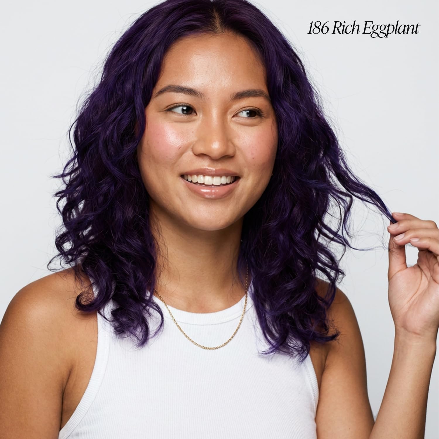 A smiling woman with shoulder-length wavy hair in Creative Image Adore #186 Rich Eggplant color, touching a strand.